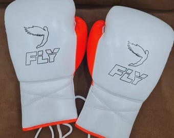 May include: A pair of white boxing gloves with orange accents. Each glove features the word "FLY" in a bold font, along with a stylized bird graphic. The gloves have white laces and are designed for boxing or other combat sports.