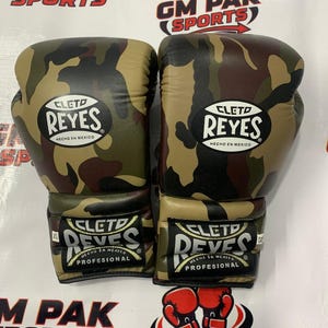 May include: Pair of camouflage boxing gloves with the "CLETO REYES" logo. The gloves are in shades of green, brown, and black. The text "HECHO EN MEXICO" is also visible. The gloves are labeled "PROFESIONAL".