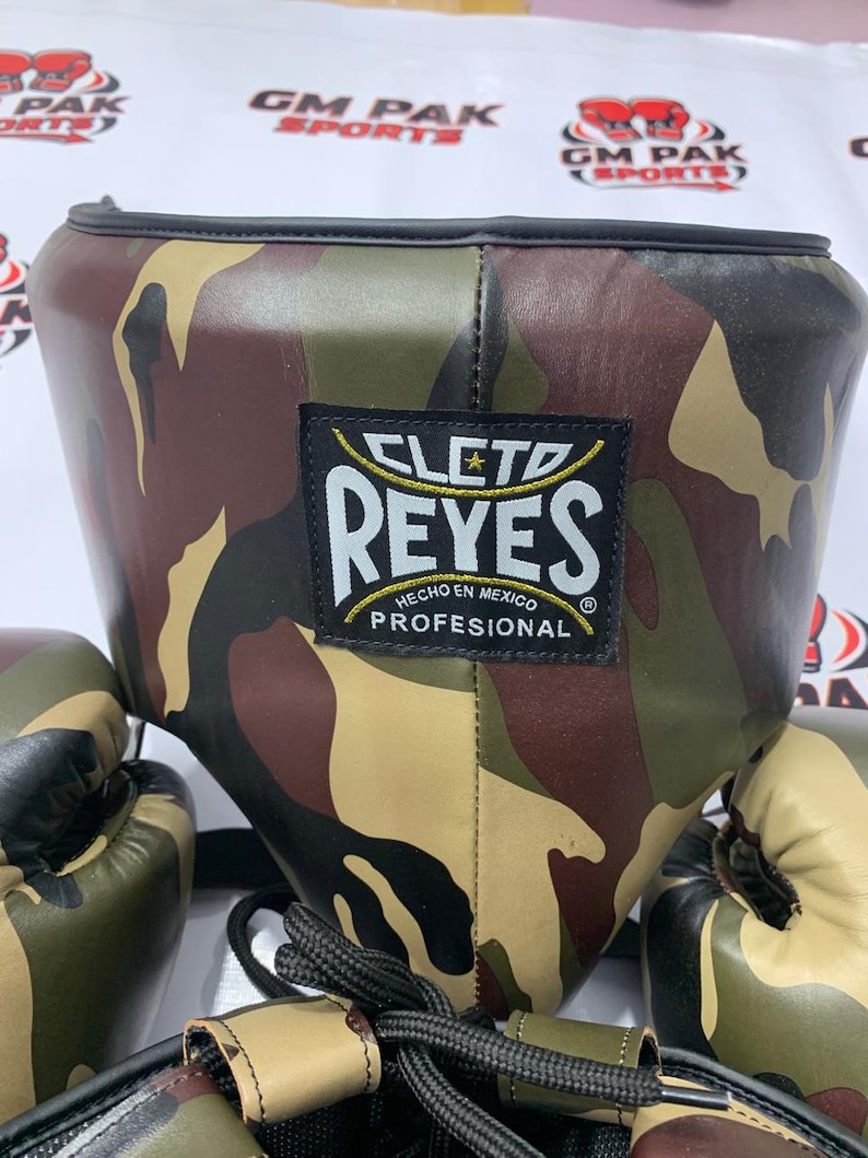 May include: Camouflage-patterned boxing headgear and gloves. The headgear features a black trim and a label that reads "CLETO REYES HECHO EN MEXICO PROFESIONAL". The gloves and headgear have a brown, green, and tan camouflage design.