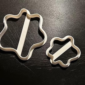May include: Two white plastic cookie cutters. The larger cutter has a wavy, shield-like shape with a diagonal bar across the center. The smaller cutter mirrors the design. Both cutters are on a dark surface.