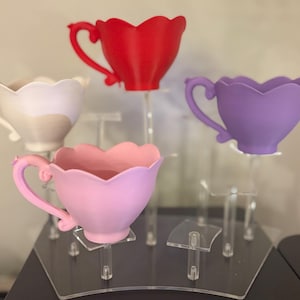 May include: Four colorful teacups in red, white, pink, and purple, each with a unique handle design and scalloped rim. The teacups are displayed on clear acrylic stands, showcasing their vibrant colors and intricate details. A charming and whimsical display.