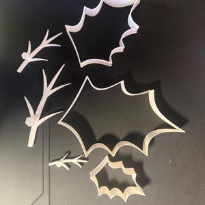 May include: A set of white plastic cookie cutters consisting of three holly leaf shapes in varying sizes and three branch-like twig shapes. The cutters feature sharp, defined edges for cutting dough into specific botanical silhouettes.
