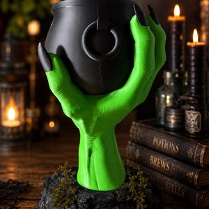 May include: A black cauldron held by a bright green hand with long black fingernails. The hand is attached to a textured black base. The cauldron features a small ring detail on its side. The design is upright and stands on a flat surface.