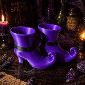 Peut inclure: Deux bottes de sorcière violettes avec des lanières noires et des bouts recourbés. Elles sont posées sur une surface en bois sombre, avec une bougie allumée, un livre intitulé "Book of Spells" et d'autres objets sur le thème de la sorcellerie.