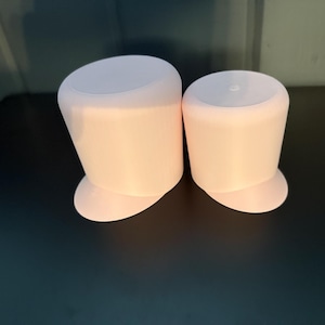 May include: Two light pink, cylindrical objects with a rounded base and a flat top. The objects are similar in shape and size, with one slightly smaller than the other. They are placed on a dark surface.