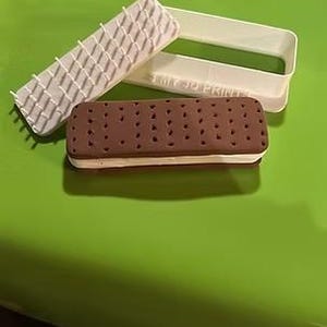 May include: A chocolate ice cream sandwich with a white filling, alongside a rectangular cookie cutter and a textured press. The sandwich has a brown exterior with small, evenly spaced dots. The cookie cutter is white.