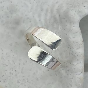 May include: A silver-colored wrap ring with a textured surface. The ring has an open design, with two curved bands that overlap. The ring is displayed on a white, textured surface.