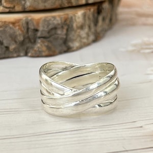 Puede incluir: Un anillo plateado con un diseño tejido. El anillo presenta múltiples bandas que se cruzan, creando una apariencia texturizada y en capas. El anillo se muestra sobre una superficie de madera de color claro.