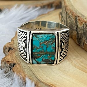 May include: A silver ring with a rectangular turquoise stone. The ring has decorative side panels with a feather design. The turquoise stone has a mix of blue and brown colors. The ring is displayed on a wooden surface.