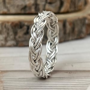 May include: A silver-colored braided ring. The ring has a woven design, with multiple strands intertwined to create a textured appearance. The ring is displayed on a light-colored wooden surface, with a blurred wooden background.