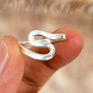 Könnte beinhalten: Ein silberfarbener Ring mit einem einzigartigen, offenen Design. Der Ring hat eine fließende, abstrakte Form. Der Ring wird zwischen zwei Fingern gehalten, was seine polierte Oberfläche und seinen modernen Stil hervorhebt.