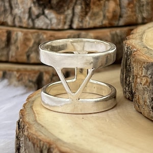 May include: A silver ring with a distinctive geometric design. The ring has two bands linked by a triangular structure. It's displayed on a wooden surface, highlighting its modern, minimalist style. This ring is a statement accessory.