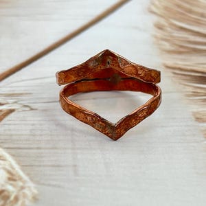 Può includere: Un anello color rame con un design a forma di V. L'anello ha una superficie strutturata e un aspetto a doppia fascia. Il gioiello è posto su uno sfondo neutro con una piuma.