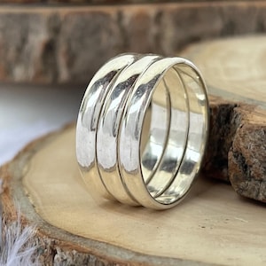 Puede incluir: Un conjunto de tres anillos plateados, cada uno con una banda lisa y redondeada. Los anillos se exhiben sobre una superficie de madera con un borde natural y rústico. Los anillos son reflectantes.