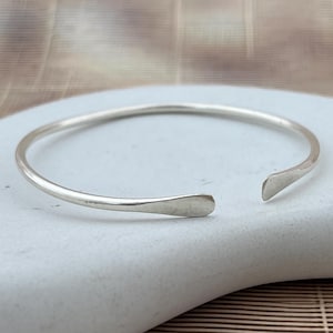 May include: A silver-colored open cuff bracelet. The bracelet has a simple, minimalist design with slightly flattened ends. The open design allows for easy wear. The bracelet is displayed on a light-colored stone surface.