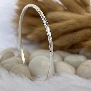 Puede incluir: Un brazalete de plata martillada. El brazalete tiene una superficie texturizada y un diseño sencillo. El fondo presenta piedras blancas y un fondo borroso de tonos neutros.