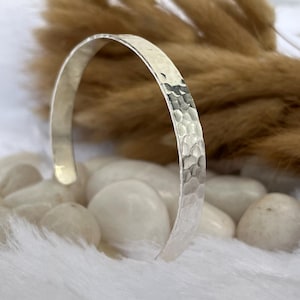 May include: A silver cuff bracelet with a hammered texture. The bracelet is open and has a simple, elegant design. The background features white stones and a blurred arrangement of dried plants.