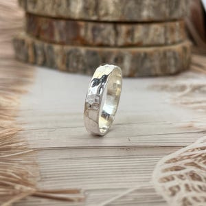 May include: A silver ring with a hammered texture. The ring is round and has a shiny, reflective surface. The ring is displayed on a light-colored wooden surface.