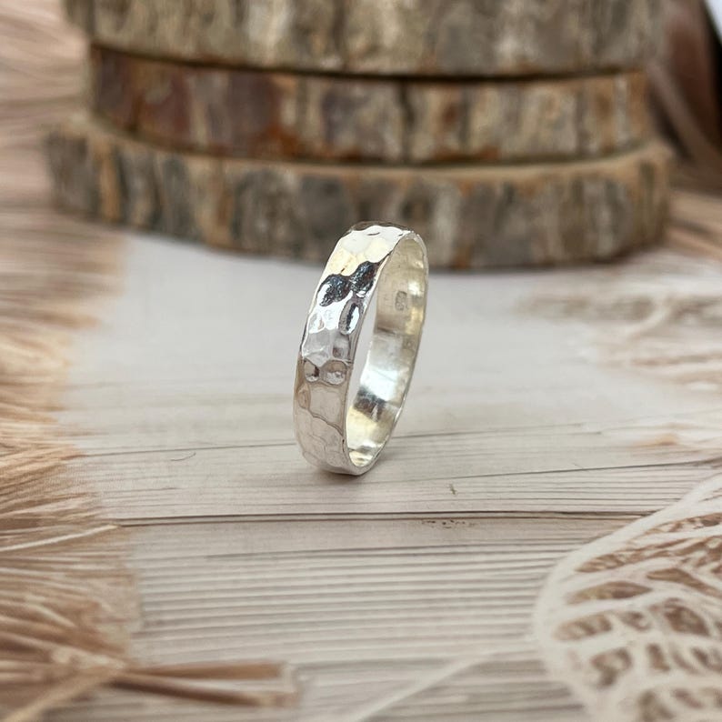 May include: A silver ring with a hammered texture. The ring is round and has a shiny, reflective surface. The ring is displayed on a light-colored wooden surface.
