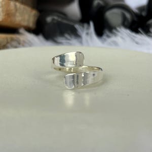 May include: A silver-colored ring with a hammered texture. The ring has an open design, with two flat, rectangular ends that overlap. The ring is set against a neutral background, highlighting its simple, modern style.