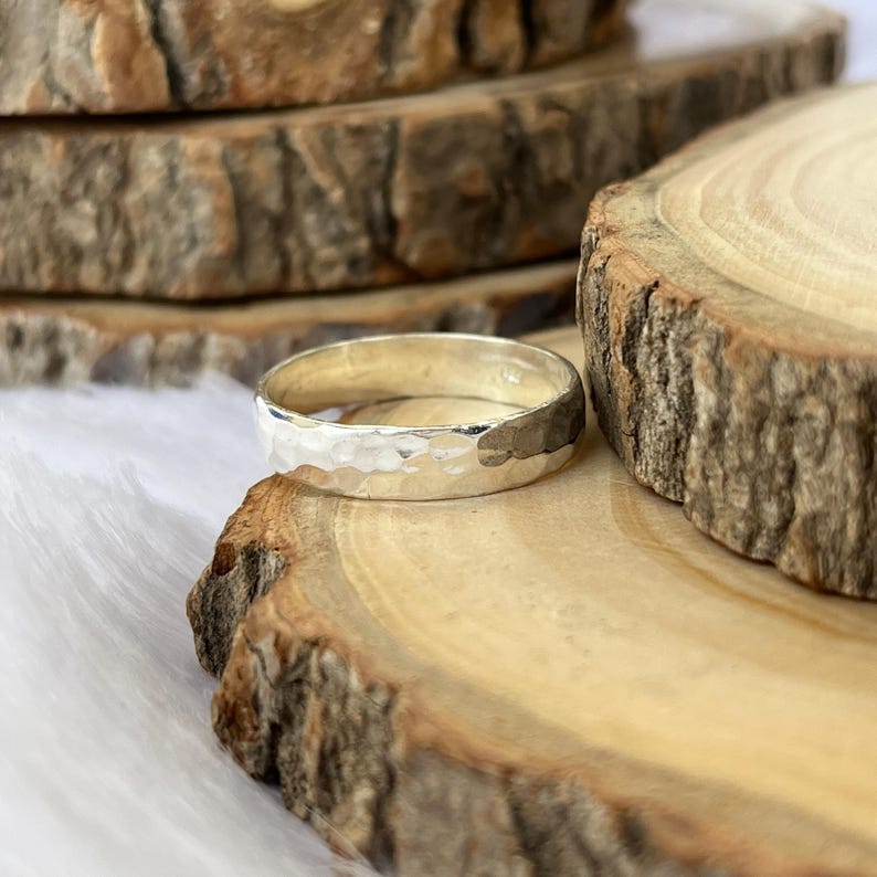 May include: A silver ring with a hammered texture. The ring is displayed on a wooden surface, showcasing its metallic shine. The ring's design features a textured surface, adding a unique visual appeal to the jewelry.