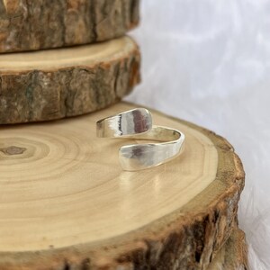 May include: A silver-colored wrap ring with a hammered texture. The ring has an open design, with the ends overlapping. The ring is displayed on a wooden surface with a natural, rustic appearance.