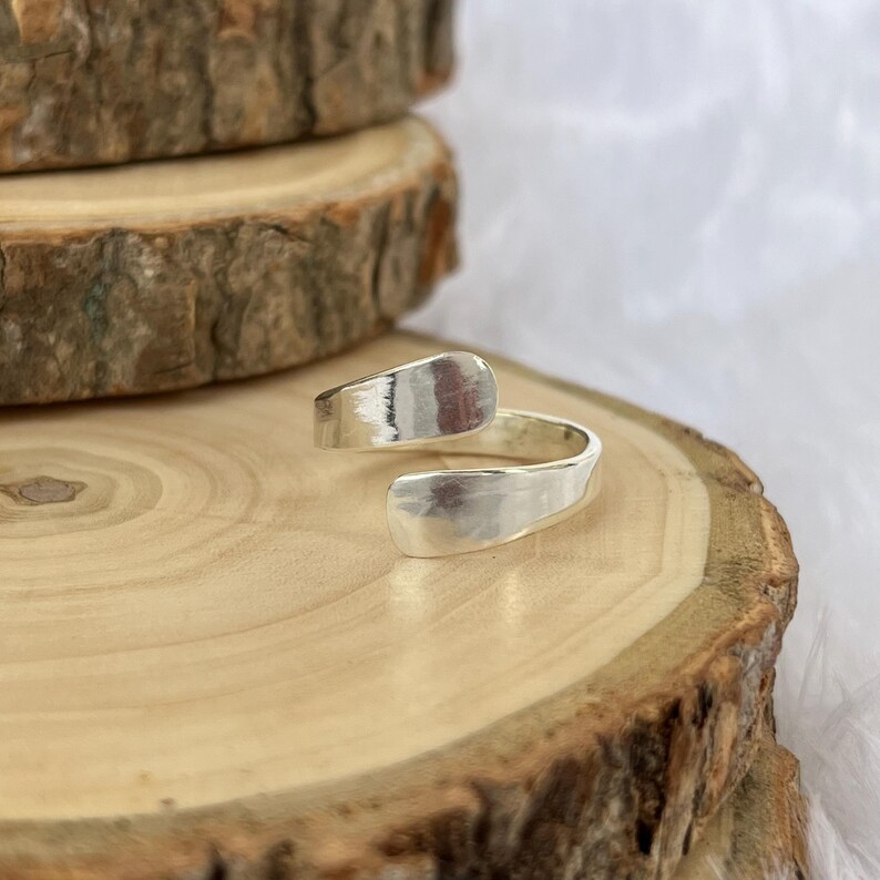 May include: A silver-colored wrap ring with a hammered texture. The ring has an open design, with the ends overlapping. The ring is displayed on a wooden surface with a natural, rustic appearance.