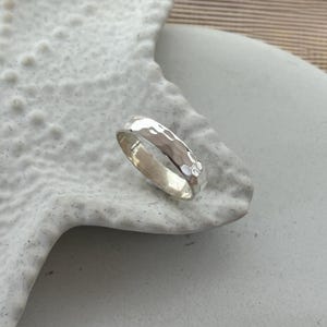 May include: A hammered silver ring with a textured surface. The ring is a band style, with a series of small indentations across the surface. The ring is displayed on a white, textured surface.