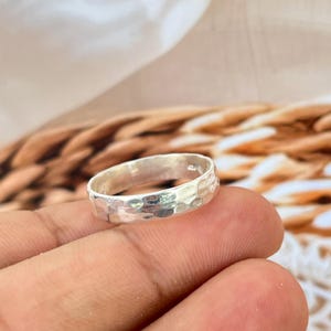 May include: A silver ring with a hammered texture. The ring is a simple band design, with a textured surface that catches the light. The ring is held in a person's hand, with a blurred background.