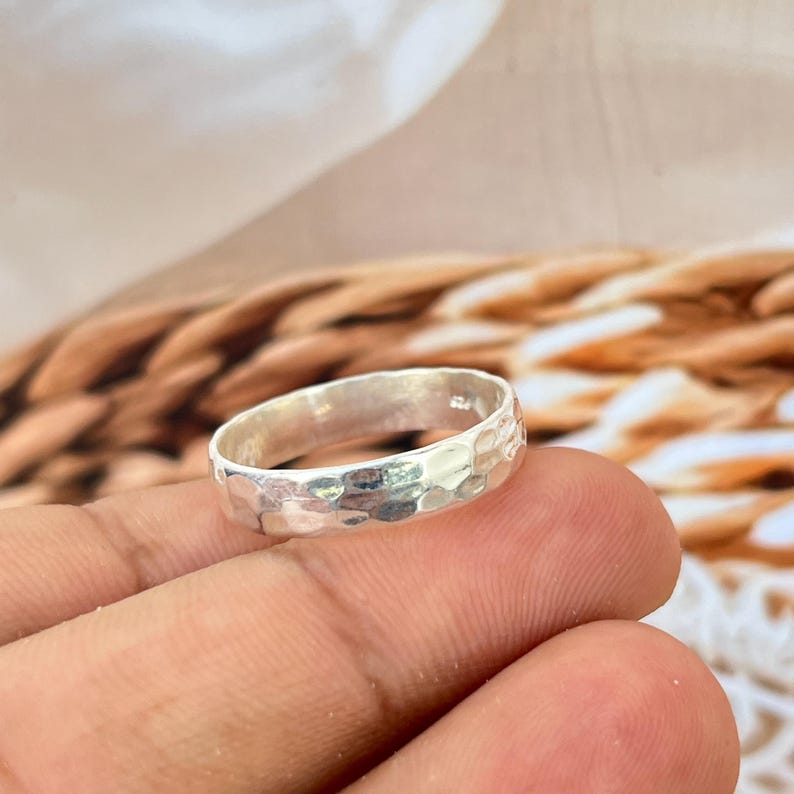 May include: A silver ring with a hammered texture. The ring is a simple band design, with a textured surface that catches the light. The ring is held in a person's hand, with a blurred background.