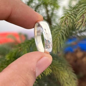 May include: A hammered silver ring held between fingers. The ring has a textured surface with a series of small indentations. The ring is a simple band design, reflecting light. The background is blurred, with green foliage.