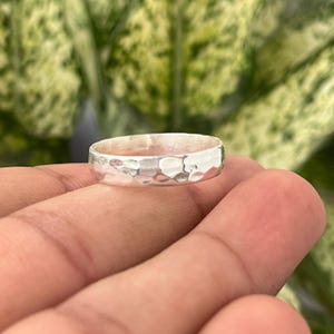 May include: A silver ring with a hammered texture. The ring is round and has a textured surface. The ring is held in a hand, with a blurred green and yellow background.