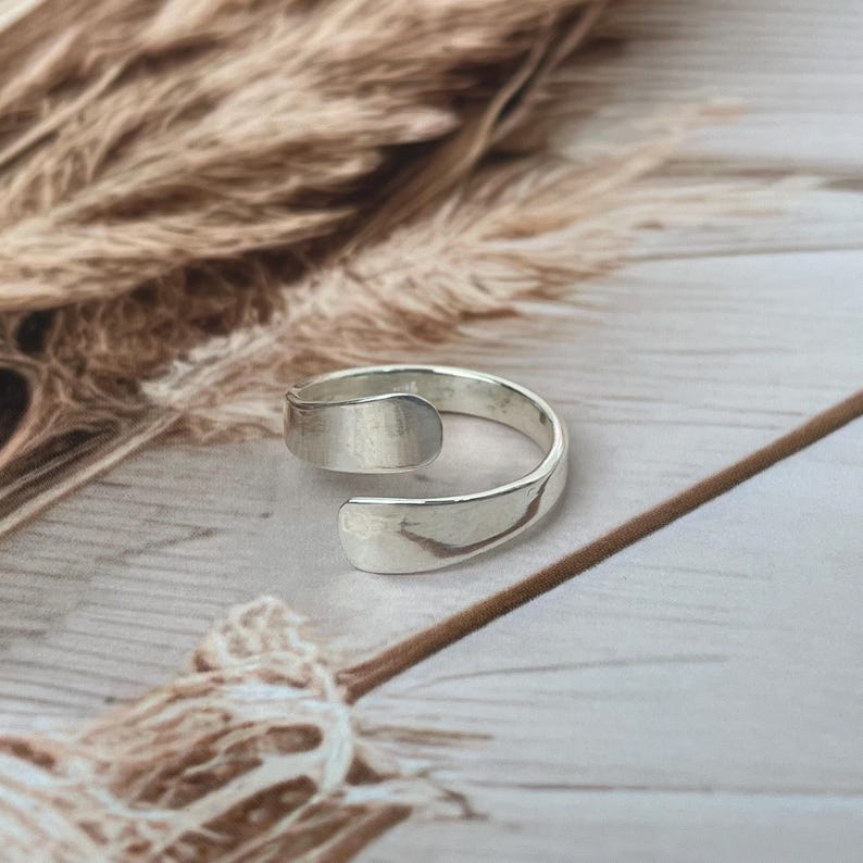 May include: A silver-colored, open-ended ring. The ring has a smooth, polished finish and a simple, minimalist design. The ring is shown on a light-colored, textured surface with dried plants in the background.