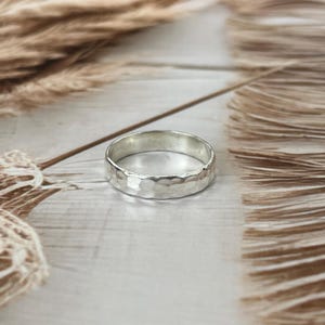 May include: A silver ring with a hammered texture. The ring is round and has a textured surface. The ring is set against a neutral background with dried plants.