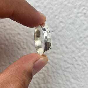 May include: A silver ring with a hammered texture. The ring is held between two fingers, showcasing its reflective surface and the detailed, irregular pattern created by the hammering process. The ring's design is simple yet eye-catching.