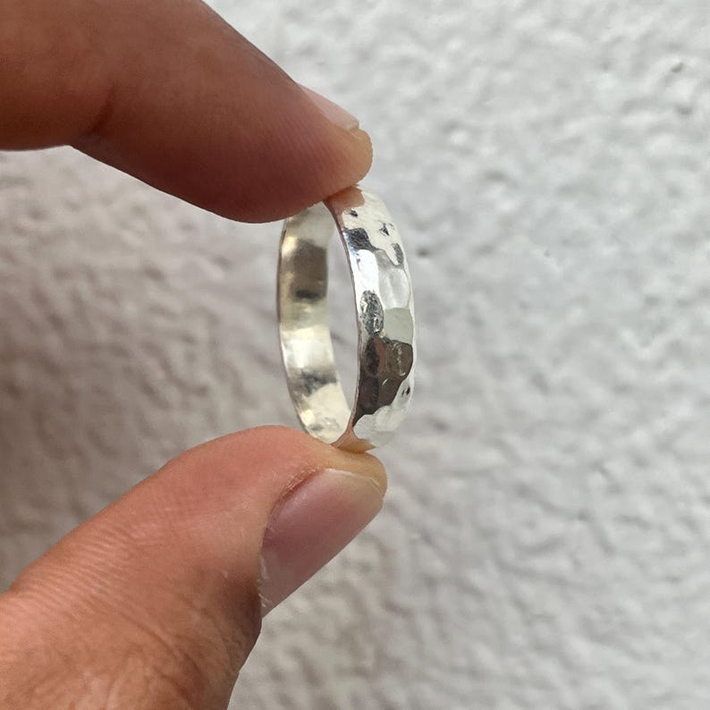 May include: A silver ring with a hammered texture. The ring is held between two fingers, showcasing its reflective surface and the detailed, irregular pattern created by the hammering process. The ring's design is simple yet eye-catching.