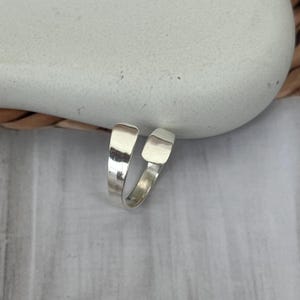May include: A silver-colored ring with a modern design. The ring features a split band with two rectangular ends that curve around. The ring is shown on a light gray surface.