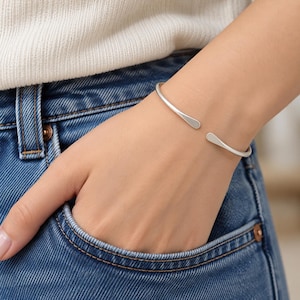 May include: A silver-toned open cuff bracelet with a smooth, matte finish and rounded, tapered ends. The bracelet features a simple, minimalist design that wraps around the wrist, leaving a small gap between the two ends of the metal band.