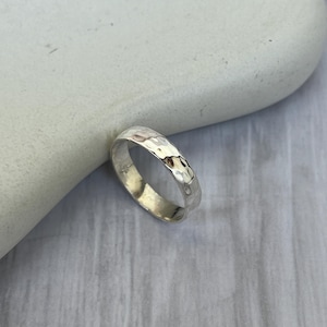 May include: A silver ring with a hammered texture. The ring is round and has a slightly irregular surface, reflecting light. The ring is displayed on a light-colored, textured surface. This ring is a piece of jewelry.