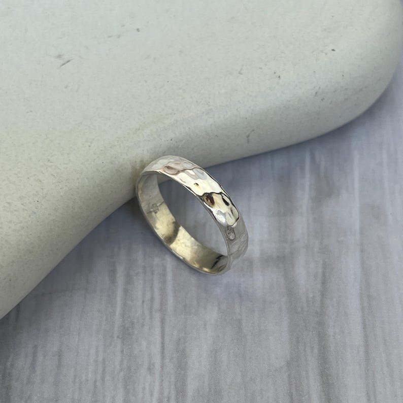 May include: A silver ring with a hammered texture. The ring is round and has a slightly irregular surface, reflecting light. The ring is displayed on a light-colored, textured surface. This ring is a piece of jewelry.
