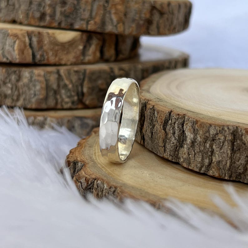 May include: A silver ring with a hammered texture. The ring is displayed on a wooden slice, with other wooden slices in the background. The ring has a shiny, reflective surface.