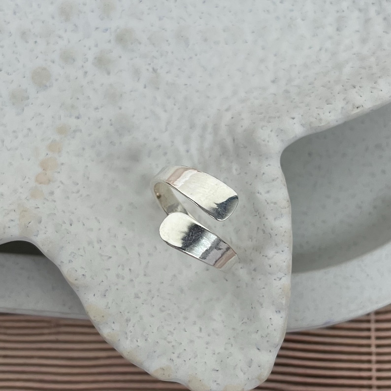 May include: A silver-colored wrap ring with a textured surface. The ring has an open design, with two curved bands that overlap. The ring is displayed on a white, textured surface.