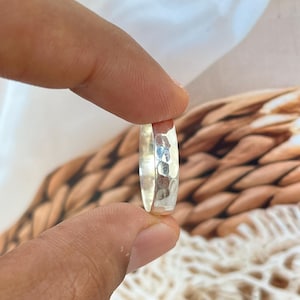 May include: A close-up of a silver ring with a hammered texture. The ring is held between two fingers, showcasing its reflective surface. The ring's design features a series of small, irregular indentations, creating a unique, textured appearance. The ring is a piece of jewelry.
