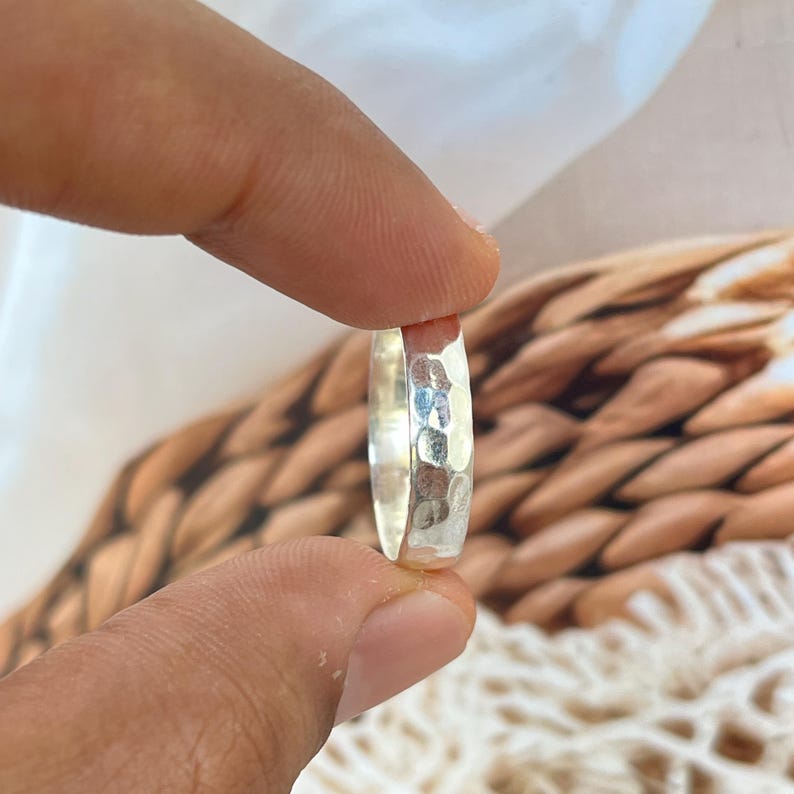 May include: A close-up of a silver ring with a hammered texture. The ring is held between two fingers, showcasing its reflective surface. The ring's design features a series of small, irregular indentations, creating a unique, textured appearance. The ring is a piece of jewelry.