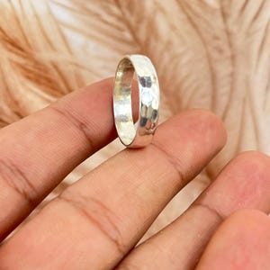 May include: A silver ring with a hammered texture. The ring is round and has a slightly domed surface. The ring is held up by a person's fingers. The background is a soft, blurred brown.