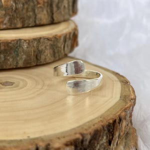 May include: A silver-colored wrap ring with a hammered texture. The ring has an open design, with the ends overlapping. The ring is displayed on a wooden surface with a natural, rustic appearance.