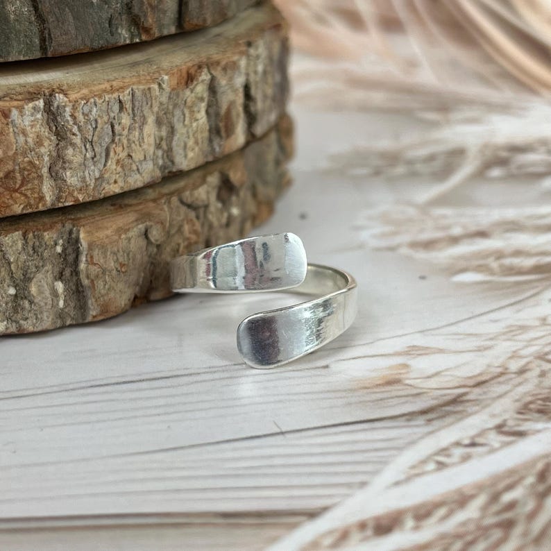 May include: A silver-colored adjustable ring with a textured finish. The ring has an open design with two rectangular ends that wrap around the finger. The ring is displayed on a light-colored surface with a natural wood background.