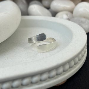 May include: A silver-colored adjustable ring with a minimalist design. The ring features a curved band that wraps around the finger, with two flat, rectangular ends. The top end has a textured finish, while the bottom end is smooth.