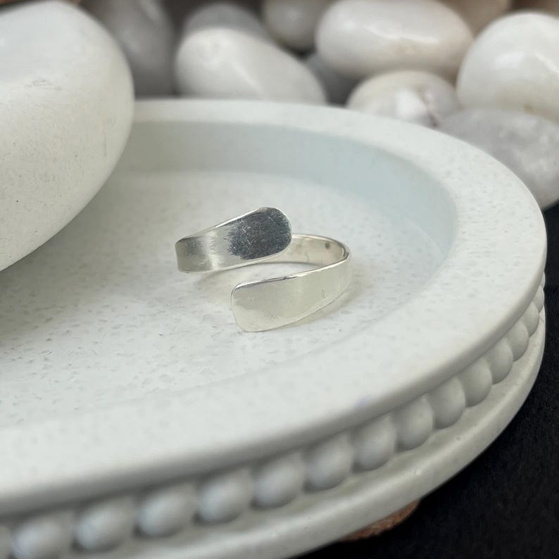 May include: A silver-colored adjustable ring with a minimalist design. The ring features a curved band that wraps around the finger, with two flat, rectangular ends. The top end has a textured finish, while the bottom end is smooth.