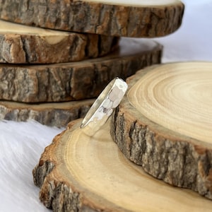 May include: A silver ring with a hammered texture, resting on a wooden surface. The ring has a geometric pattern and a shiny finish. The background features additional wooden slices, creating a rustic aesthetic. The ring is a piece of jewelry.
